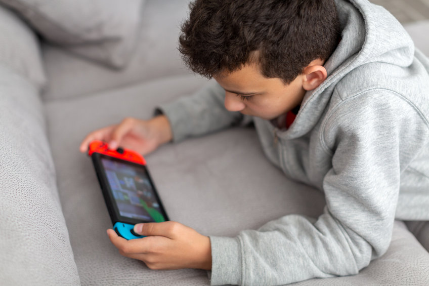 Teenager lies on a sofa and plays a game. He has a portable game console in his hands. How teens spend their vacations